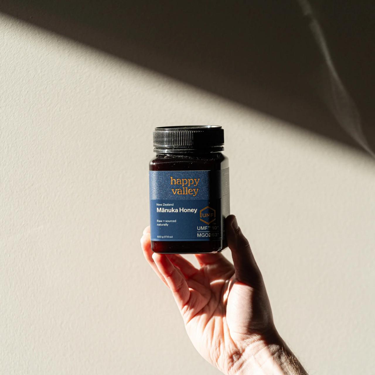 Hand holding a jar of 'Happy Valley' Manuka Honey against a neutral background